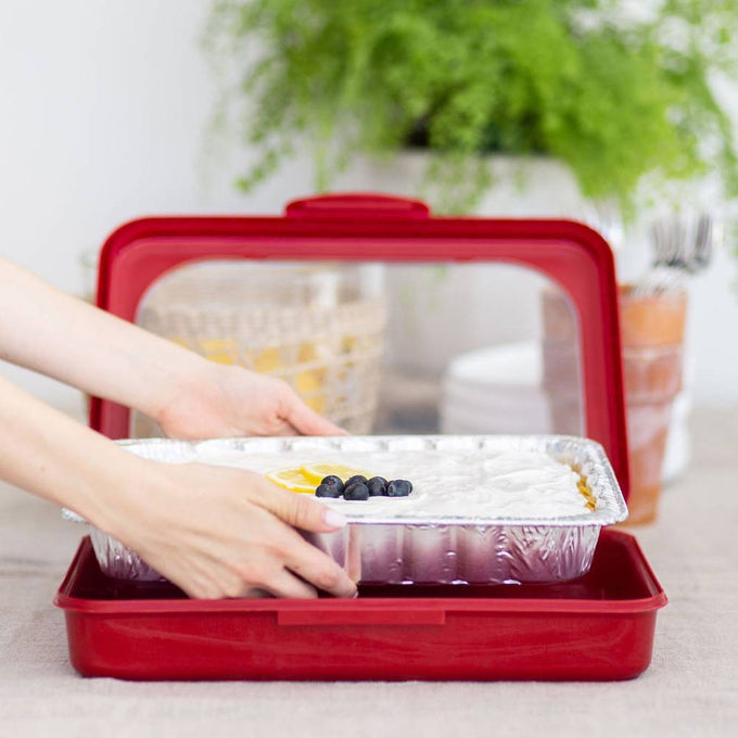 Person placing a foil pan filled with dessert topped with blueberries into a vibrant red Fancy Panz travel cover, showcasing a sturdy and stylish kitchen accessory from USA Made and Kitchen & Food collections.