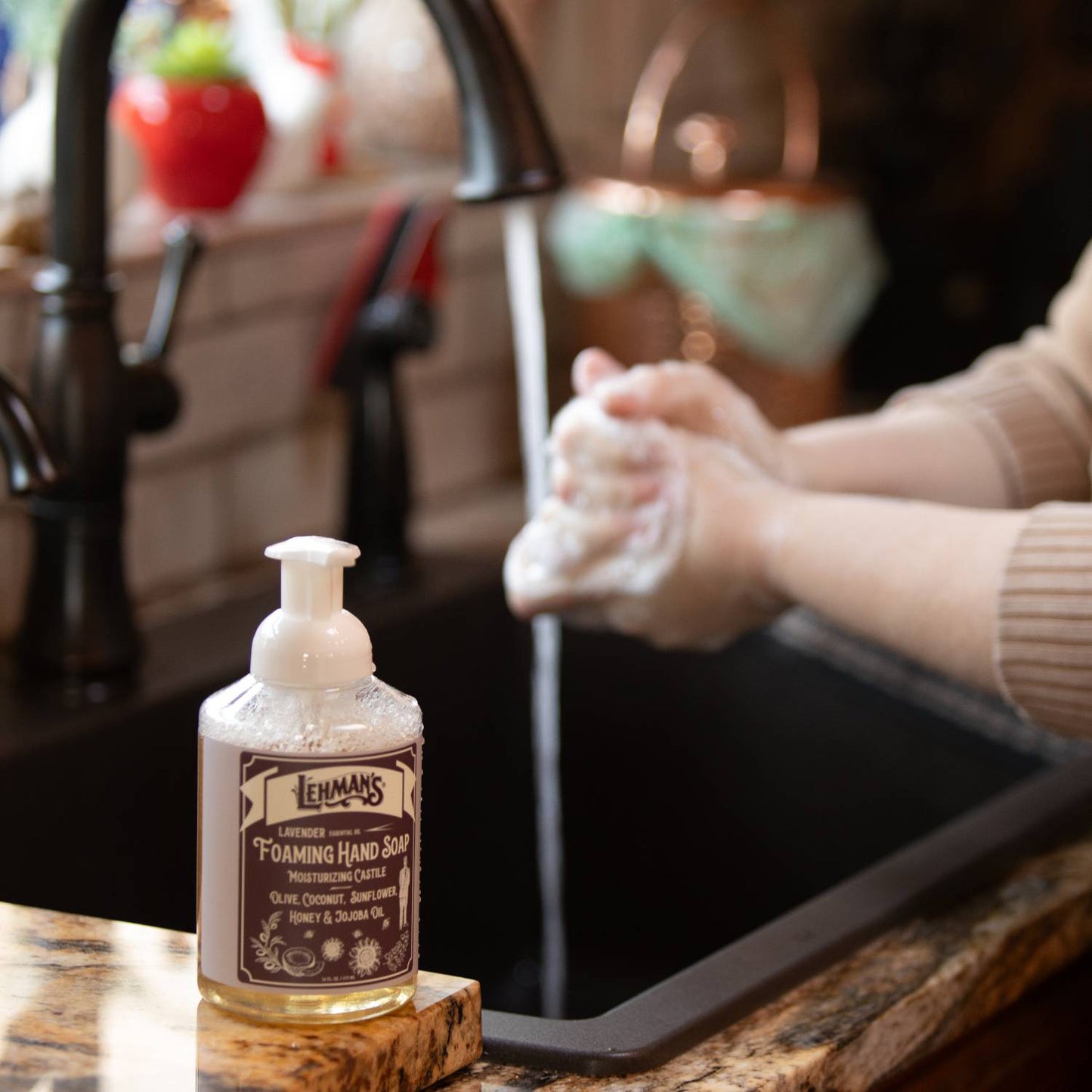 Clear pump bottle of handcrafted moisturizing foaming hand soap with natural oils and honey, made in USA, placed on a kitchen counter next to a person washing hands, ideal for Health & Wellness and Grooming collections.