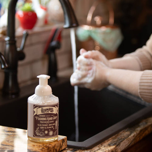 Clear pump bottle of handcrafted moisturizing foaming hand soap with natural oils and honey, made in USA, placed on a kitchen counter next to a person washing hands, ideal for Health & Wellness and Grooming collections.