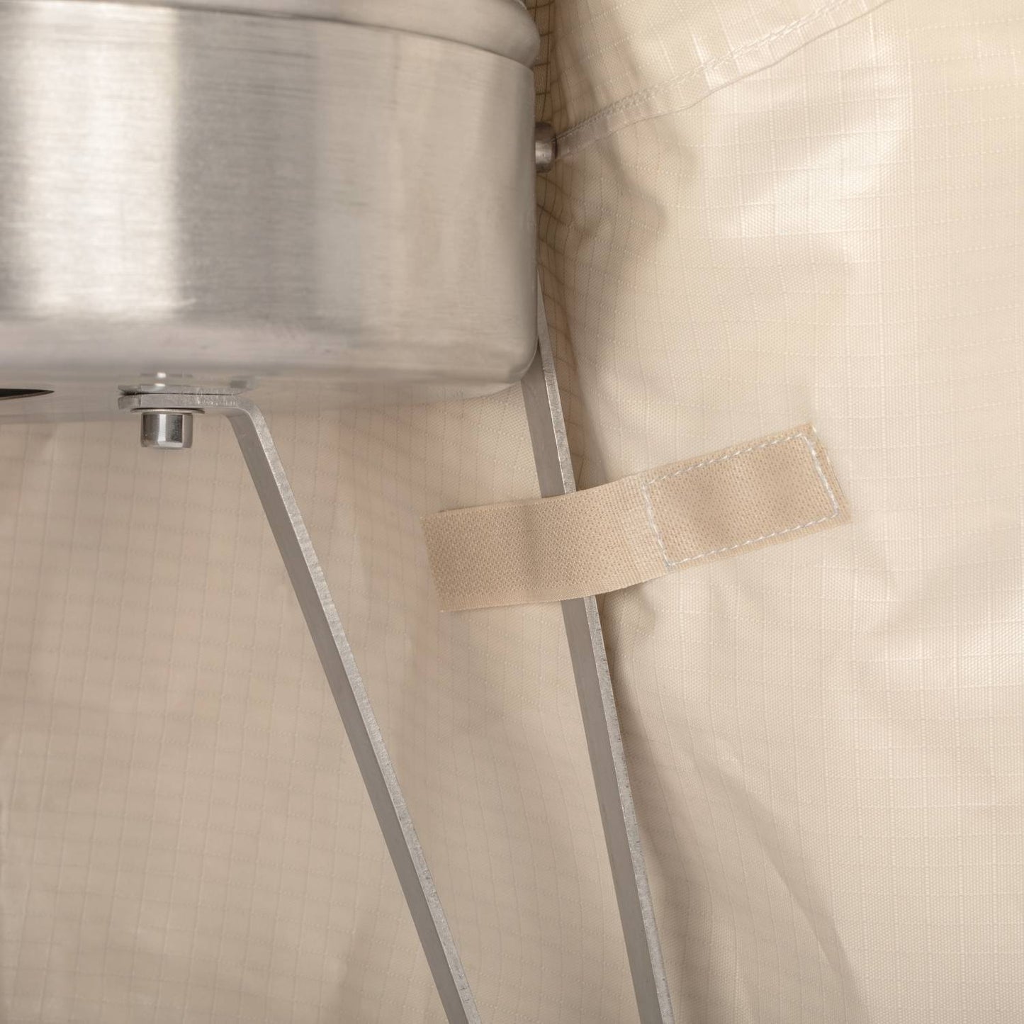 Close-up of beige ripstop fabric with a fastening strap secured beside a metal fire barrel leg, showcasing durable weatherproof protection designed for outdoor Farm, Garden & Tool and Camping use.