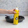 Hand holding a yellow can of Lodge seasoning spray with cast iron skillet graphic next to a black cast iron pan on a kitchen counter, suitable for Kitchen & Food, Cookware, Cast Iron collections.
