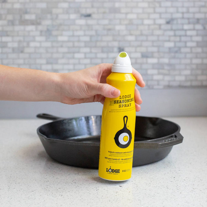 Hand holding a yellow can of Lodge seasoning spray with cast iron skillet graphic next to a black cast iron pan on a kitchen counter, suitable for Kitchen & Food, Cookware, Cast Iron collections.