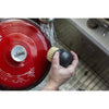 Hand holding a round brush with stiff bristles under running water near a red enameled cast iron pot lid, ideal for maintaining cookware in Kitchen & Food and Cast Iron collections.