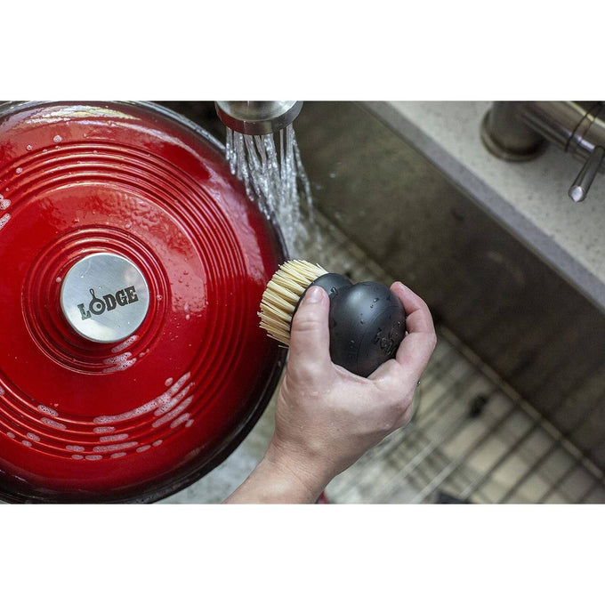 Hand holding a round brush with stiff bristles under running water near a red enameled cast iron pot lid, ideal for maintaining cookware in Kitchen & Food and Cast Iron collections.