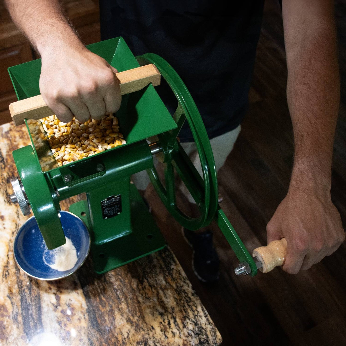 Person using a green manual grain mill with wooden handle to grind whole corn kernels into flour, set on a granite countertop, suitable for homesteading and emergency supplies in Farm, Garden & Tool and Kitchen & Food collections.
