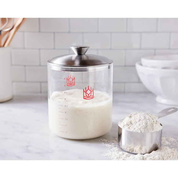 Clear borosilicate glass crock with stainless steel lid holding sourdough starter beside a metal measuring cup filled with flour, perfect for Kitchen & Food, Baking Supplies, and Cookware collections.