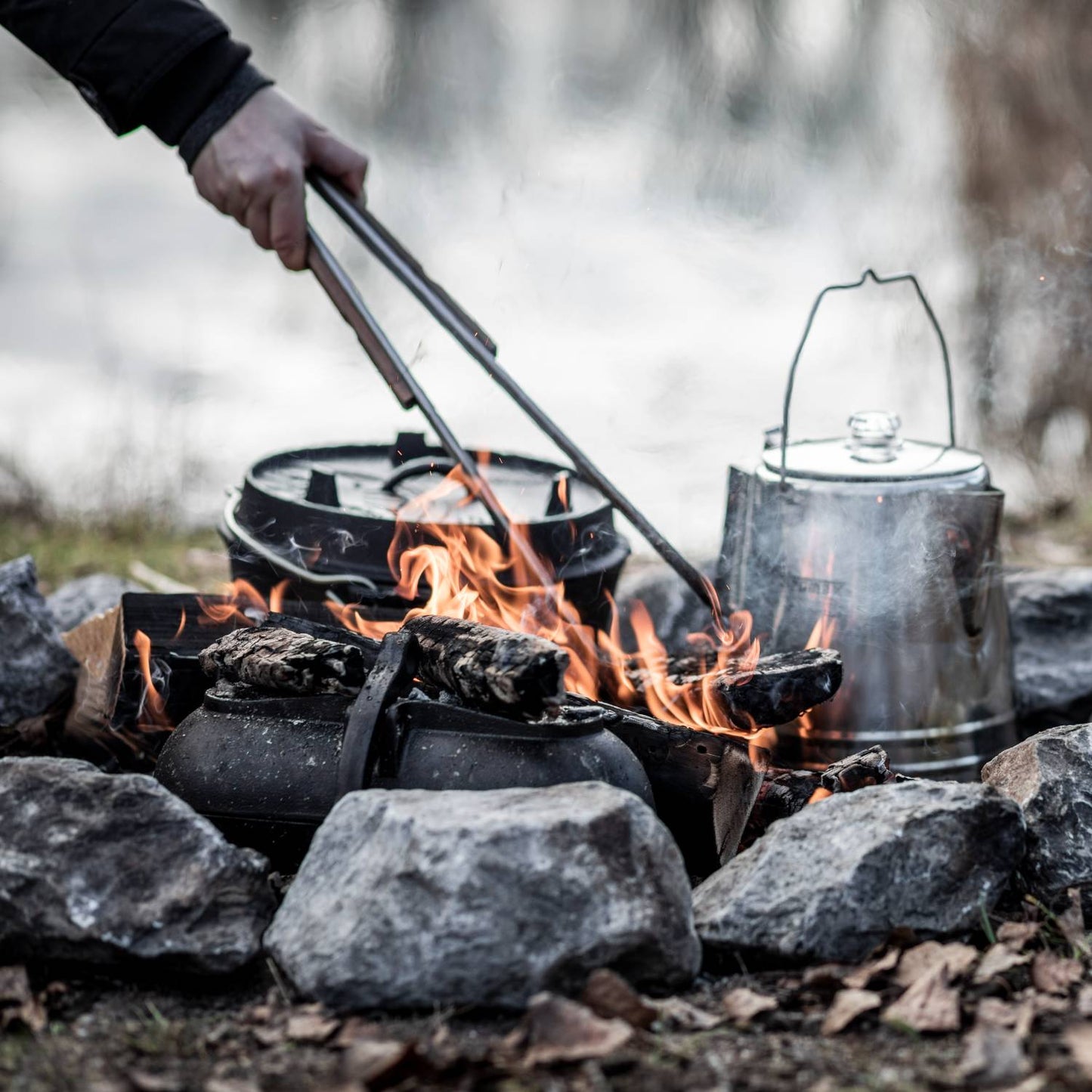 Brushed stainless steel tongs with wooden grips being used to safely handle hot coals over an outdoor campfire setup with cast iron pots and a metal kettle, ideal for grilling, camping, and outdoor cooking in Farm, Garden & Tool and Camping collections.