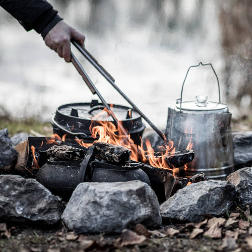 Brushed stainless steel tongs with wooden grips being used to safely handle hot coals over an outdoor campfire setup with cast iron pots and a metal kettle, ideal for grilling, camping, and outdoor cooking in Farm, Garden & Tool and Camping collections.