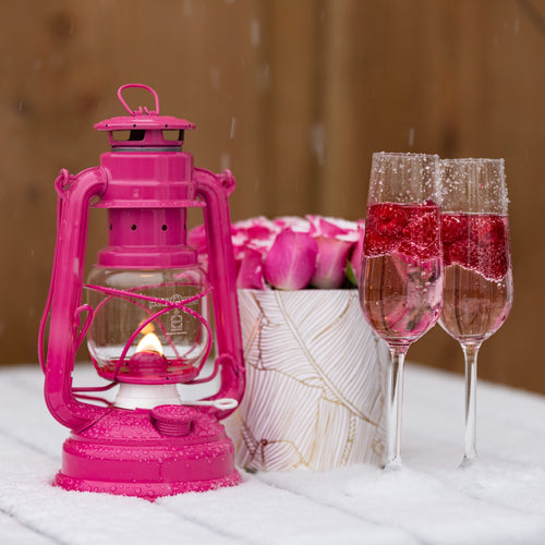Handcrafted bright pink galvanized oil lantern with thermal heat-resistant globe glowing warmly beside a floral arrangement and two sparkling rose wine glasses, ideal for Camping, Lamps & Lighting, and Emergency Supplies collections.