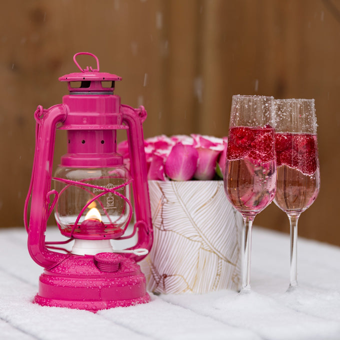 Handcrafted bright pink galvanized oil lantern with thermal heat-resistant globe glowing warmly beside a floral arrangement and two sparkling rose wine glasses, ideal for Camping, Lamps & Lighting, and Emergency Supplies collections.