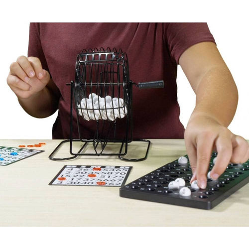 Person using a metal rotary bingo cage with white balls, marking numbers on bingo cards and organizing balls on a plastic call sheet, part of Toys, Books & Games collection.
