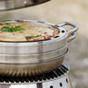 Close-up of a round stainless steel baking tray with ventilation holes and handles, holding a crispy baked pizza garnished with basil, set on an outdoor camping oven, ideal for Farm, Garden & Tool, Camping, and Best Sellers collections.