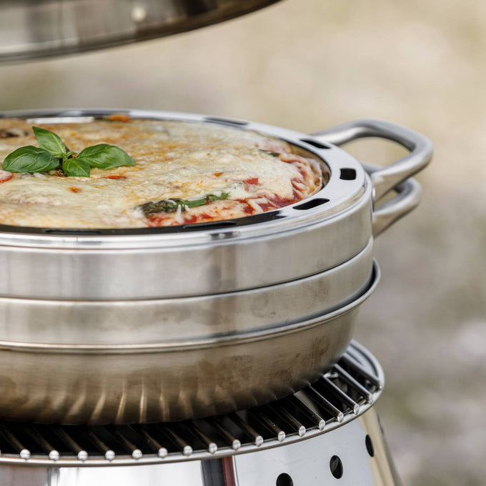 Close-up of a round stainless steel baking tray with ventilation holes and handles, holding a crispy baked pizza garnished with basil, set on an outdoor camping oven, ideal for Farm, Garden & Tool, Camping, and Best Sellers collections.