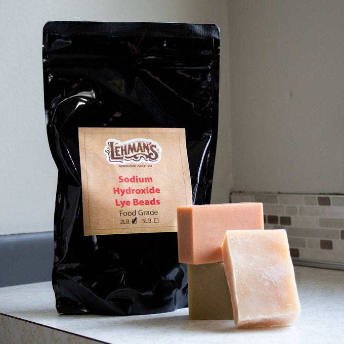 Black resealable bag of food-grade sodium hydroxide lye beads alongside three handmade soap bars, featured in Health & Wellness, Soaps and Soapmaking, and USA Made collections.