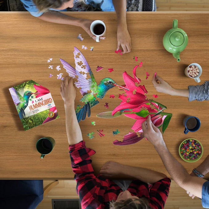 People assembling a 300-piece shaped hummingbird jigsaw puzzle with vibrant colors on a wooden table, surrounded by mugs, a teapot, and snacks, ideal for Toys, Books & Games collections.