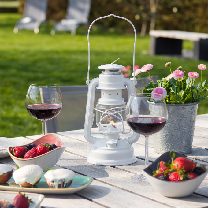 Handcrafted white galvanized oil lantern with thermal heat-resistant globe softly glowing on a light wood table surrounded by fresh berries, two glasses of red wine, and a pot of pink flowers, ideal for Camping, Lamps & Lighting, and Emergency Supplies collections.