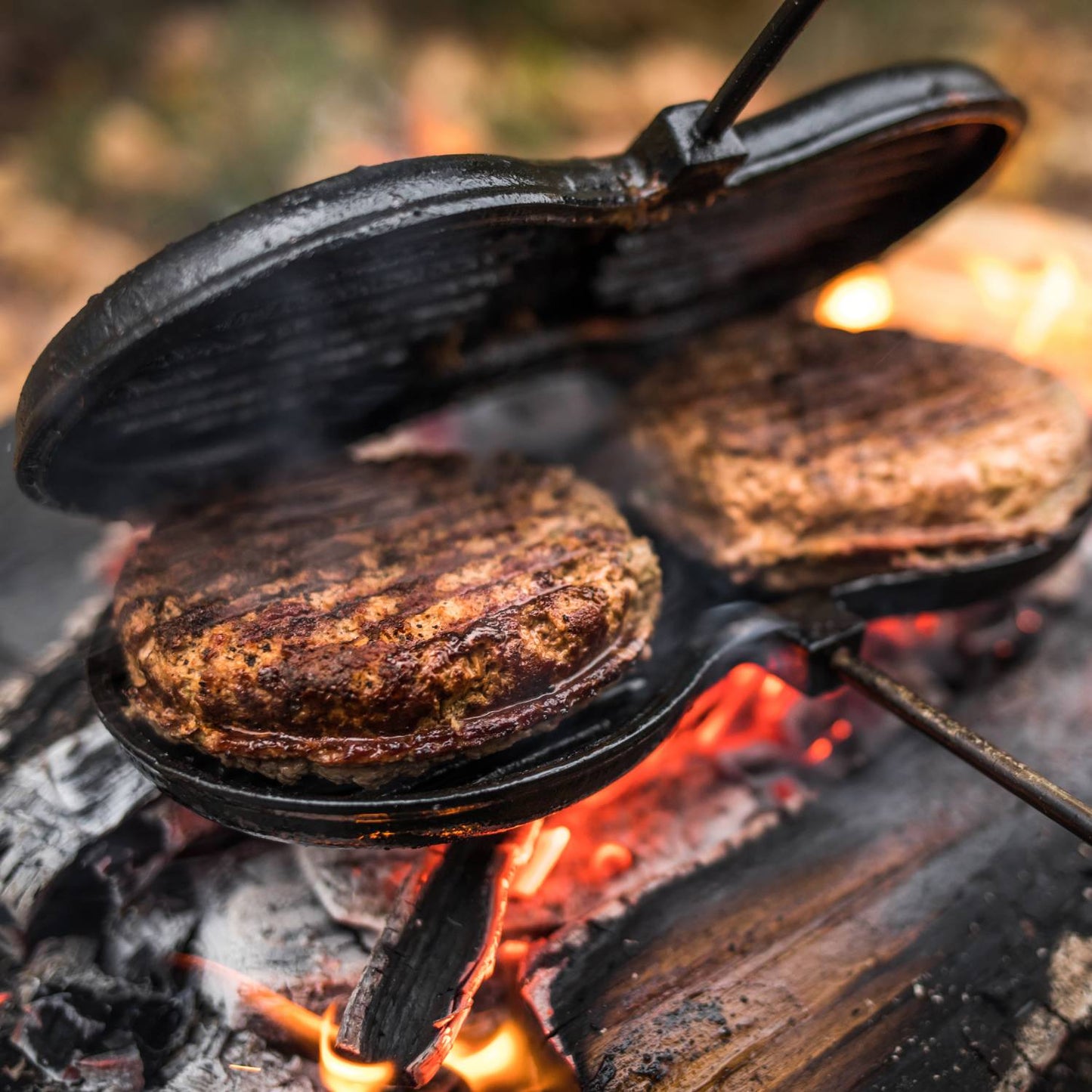 Cast iron burger iron with two round ridged molds cooking juicy patties over an open campfire, designed for outdoor use in Camping, Cookware, and Cast Iron collections.
