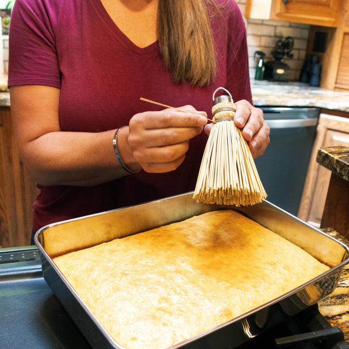 Person holding a handcrafted Amish-style broom cake tester with a metal loop, poised to poke a straw into a freshly baked sheet cake in a kitchen, featured in Kitchen & Food and USA Made collections.