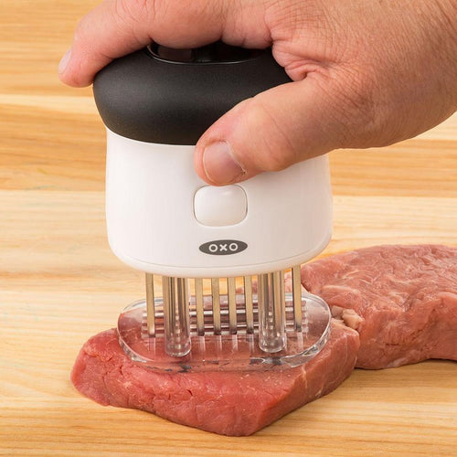 Hand pressing a white and black bladed meat tenderizer with 58 stainless steel blades into a raw cut of meat on a wooden surface, part of Kitchen & Food, Specialty Equipment, and Butchering Supplies collections.