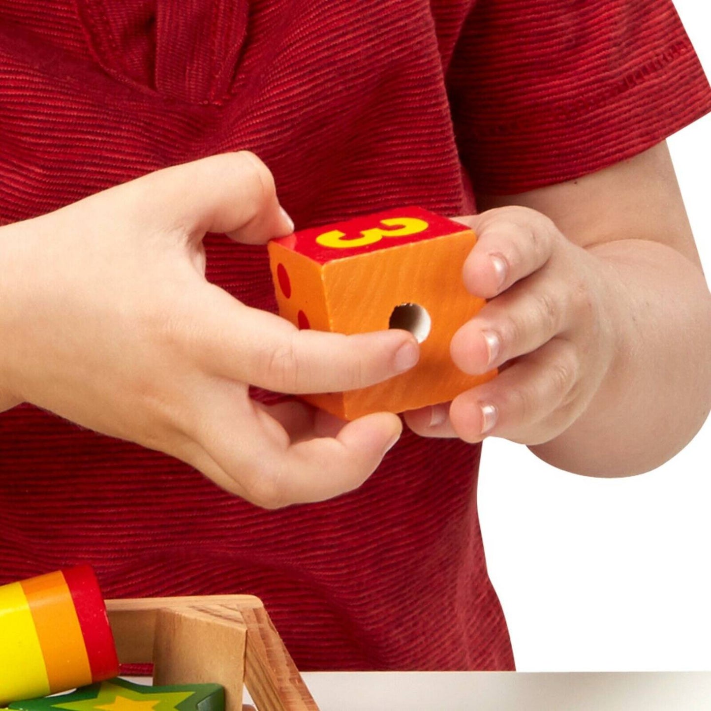 Child wearing red shirt holding a bright orange wooden lacing bead with the number 3, part of a colorful educational toy set for indoor play and fine motor skill development.