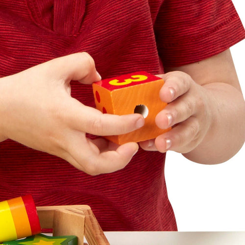Child wearing red shirt holding a bright orange wooden lacing bead with the number 3, part of a colorful educational toy set for indoor play and fine motor skill development.