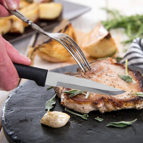 Handmade serrated steak knife with high carbon surgical stainless steel blade and solid black handle cutting a grilled pork chop on a rustic slate plate, ideal for Kitchen & Food and USA Made collections.