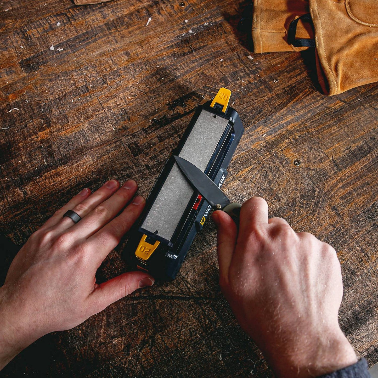 Person sharpening a knife on a benchtop sharpening system with coarse and fine abrasive surfaces, placed on a worn wooden table near a leather glove, suited for Farm, Garden & Tool and Kitchen & Food collections.
