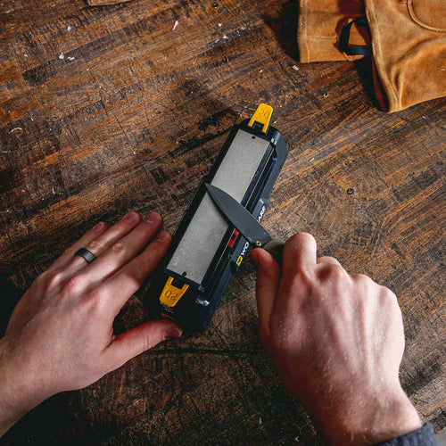 Person sharpening a knife on a benchtop sharpening system with coarse and fine abrasive surfaces, placed on a worn wooden table near a leather glove, suited for Farm, Garden & Tool and Kitchen & Food collections.
