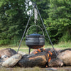Black iron tripod with adjustable chain holding a cast iron pot suspended over an open campfire surrounded by large rocks, suitable for outdoor cooking in camping and farm collections.