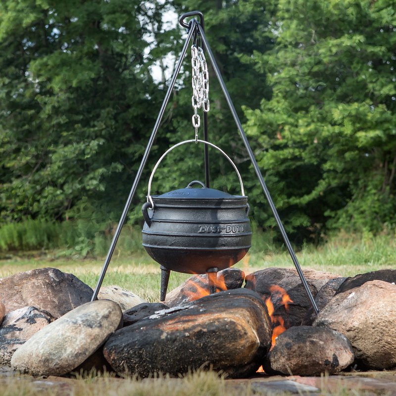 Black iron tripod with adjustable chain holding a cast iron pot suspended over an open campfire surrounded by large rocks, suitable for outdoor cooking in camping and farm collections.