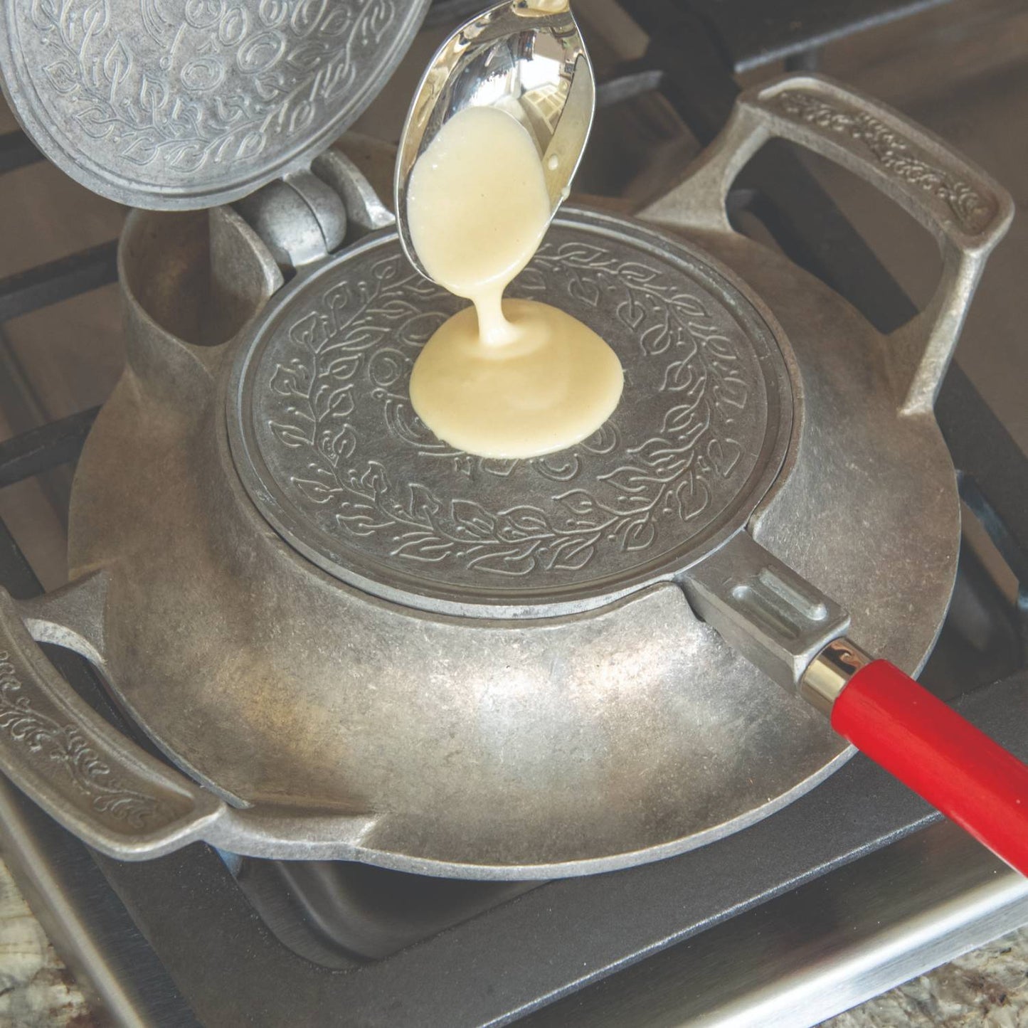 Pouring batter onto a handcrafted stovetop iron with decorative patterns and a red wooden handle, designed for making traditional cookies, featured in kitchen and cookware collections.
