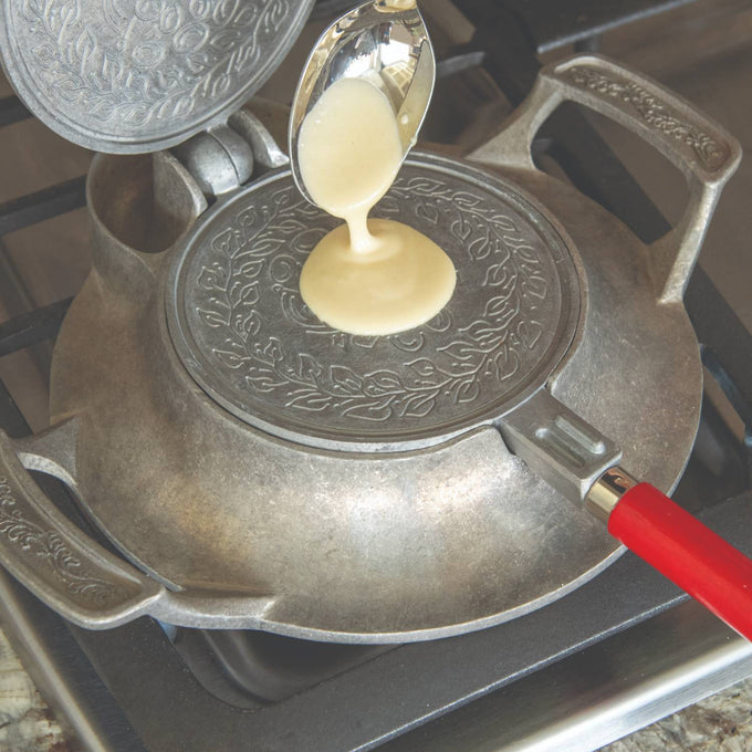Pouring batter onto a handcrafted stovetop iron with decorative patterns and a red wooden handle, designed for making traditional cookies, featured in kitchen and cookware collections.