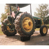Green and yellow John Deere tractor resting on a gravel path with a heavy-duty black block supporting the rear tire, surrounded by green pine trees and farmland background