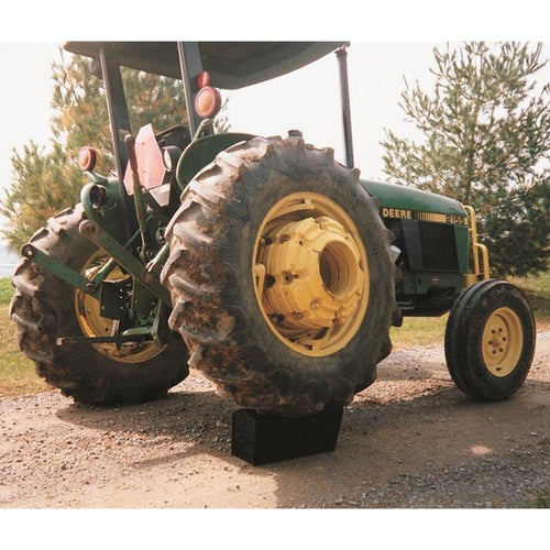 Green and yellow John Deere tractor resting on a gravel path with a heavy-duty black block supporting the rear tire, surrounded by green pine trees and farmland background