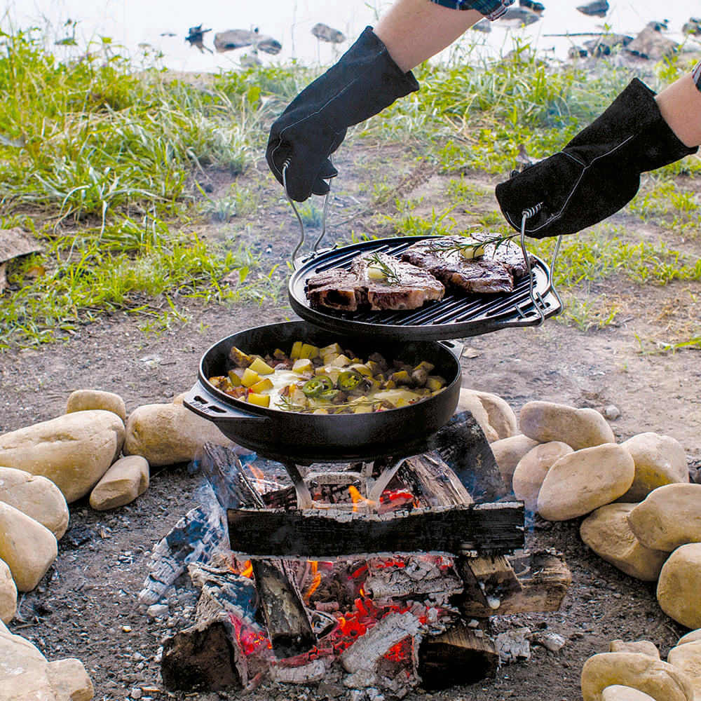 Handcrafted cast iron pan set in use over an outdoor campfire with gloves lifting a grilling grate holding steaks above a pan of cooked vegetables, perfect for camping and outdoor cooking in Kitchen & Food and Cast Iron collections.