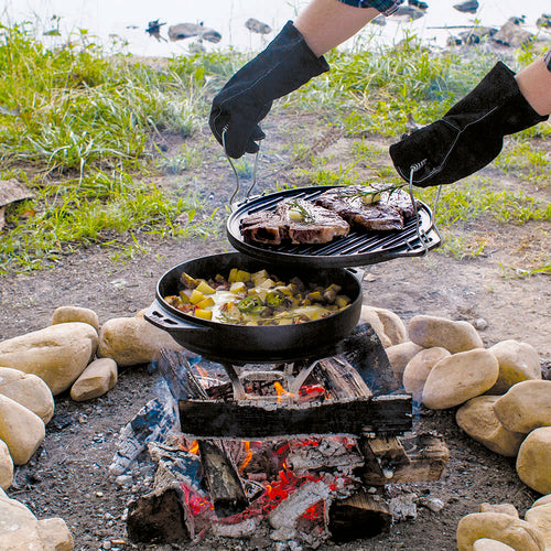 Handcrafted cast iron pan set in use over an outdoor campfire with gloves lifting a grilling grate holding steaks above a pan of cooked vegetables, perfect for camping and outdoor cooking in Kitchen & Food and Cast Iron collections.
