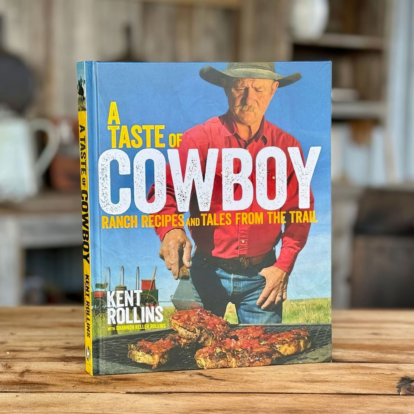 Hardcover cookbook titled "A Taste of Cowboy" featuring ranch recipes and trail stories by Kent Rollins, standing upright on wooden surface, part of Books and Cookbooks collections.