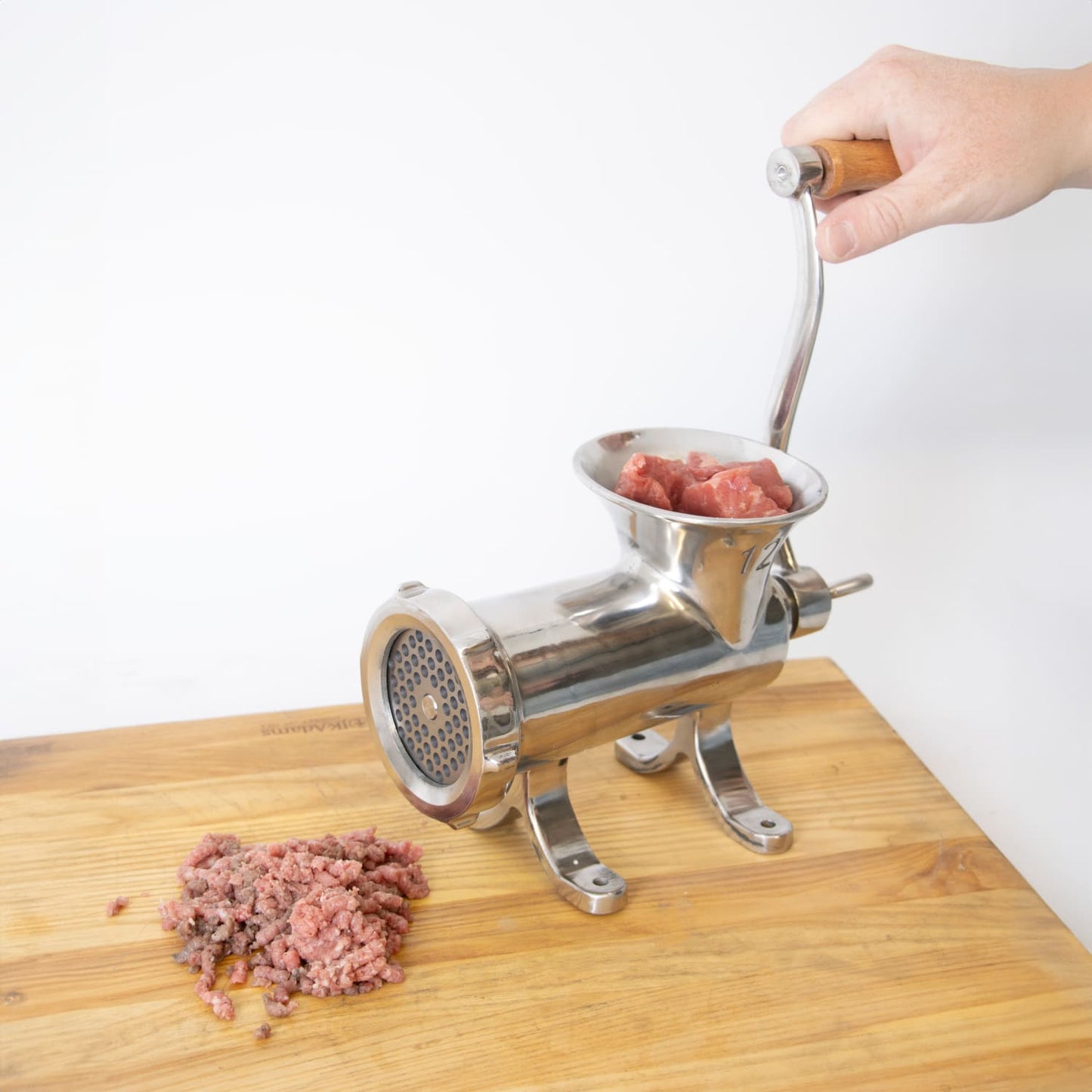 Handcrafted stainless steel mounted meat grinder with wooden handle being hand-cranked on wooden surface, processing fresh chunks of meat, featured in Kitchen & Food, Specialty Equipment, Butchering Supplies, Best Sellers, Promo Eligible collections.