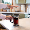 Person pressing down the plunger on a stainless steel and glass French press coffee maker with black handle in a bright kitchen, featured in Kitchen & Food, Brewing Coffee and Tea, Best Sellers, and Promo Eligible collections.