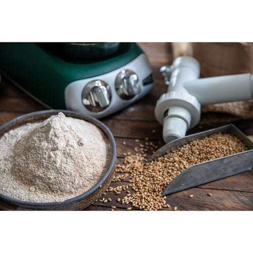 White grain mill attachment connected to a green stand mixer with freshly ground flour in a bowl and scattered wheat grains on a wooden surface, perfect for Kitchen & Food, Baking Supplies, Grain Mills, and Bread Making collections.