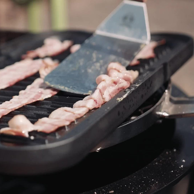 Close-up of bacon strips cooking on the ribbed side of a handcrafted nitrogen hardened cast iron griddle, with a metal spatula lifting one piece, suitable for grilling and cookware collections.