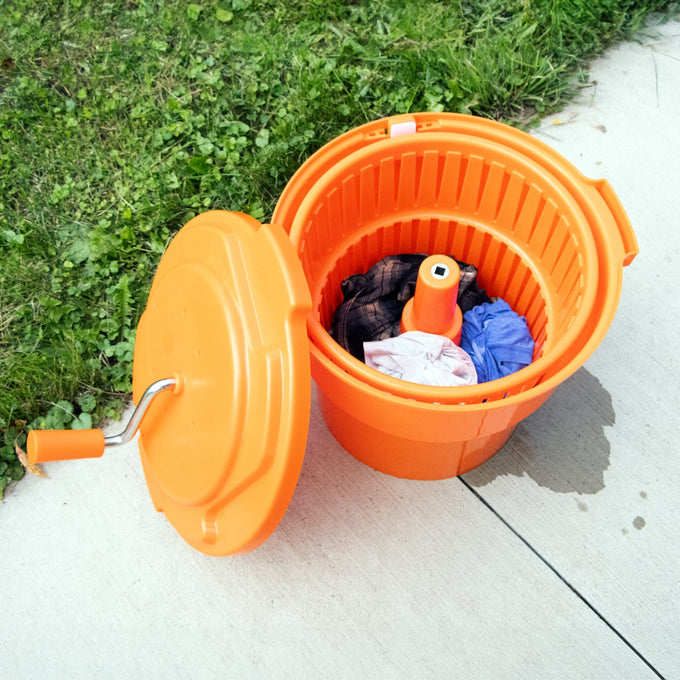 Bright orange handcrafted hand-powered laundry spinner with removable lid and crank handle, holding wet clothes on a sidewalk near grass, part of Home & Decor and Laundry Supplies collections.