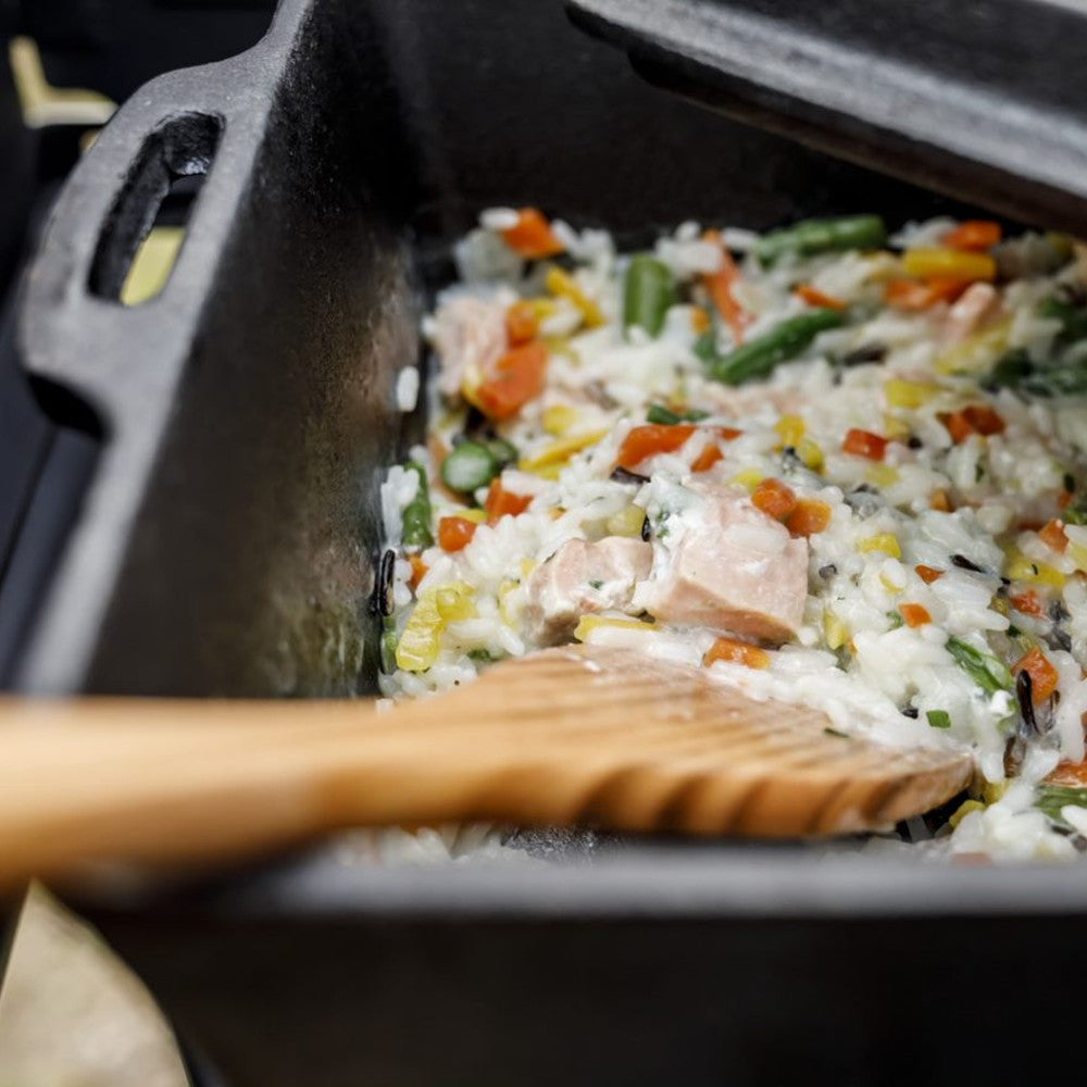 Close-up of a rustic cast iron loaf pan cooking a creamy rice dish with mixed vegetables and chunks of meat, ideal for farm, kitchen, camping, cookware, and cast iron collections.