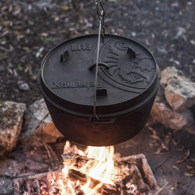 Heavy-duty cast iron Dutch oven with legs suspended over a campfire, featuring a detailed embossed lid with a sturdy wire handle, ideal for Kitchen & Food, Cookware, Cast Iron, Best Sellers, Promo Eligible collections.