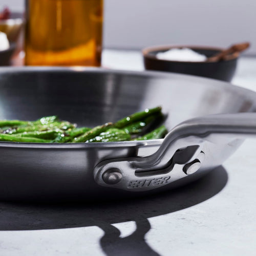 Close-up of a handcrafted fully clad stainless steel fry pan handle with the word "Eater" engraved, set against a kitchen backdrop with green vegetables cooking, highlighted for kitchen and cookware collections.