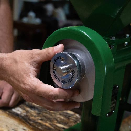 Hand adjusting the fineness setting on a sturdy green grain mill with stainless steel burrs, designed for homesteading, kitchen, and emergency food grinding needs.