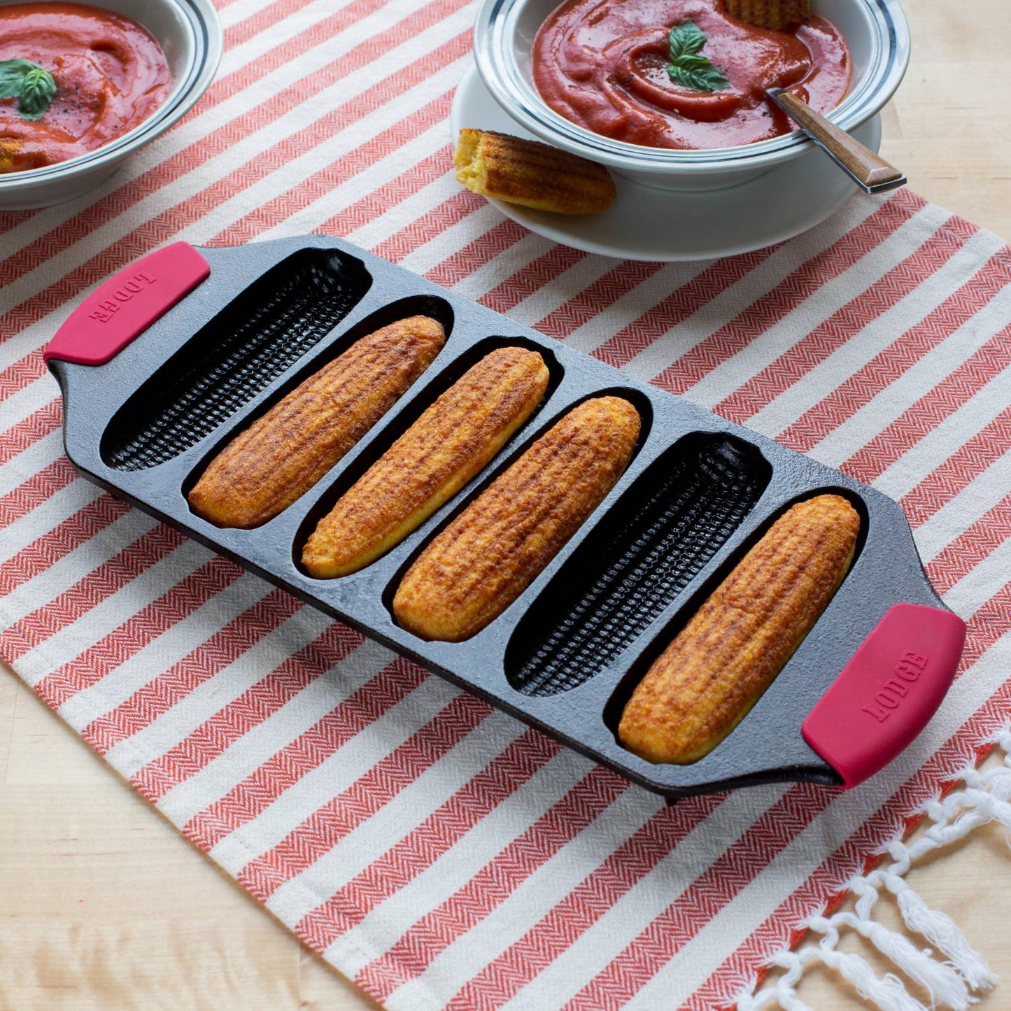 Cast iron pan with six crispy cornsticks, featuring red silicone handles, resting on a striped cloth next to bowls of tomato soup, ideal for Kitchen & Food, Cookware, Cast Iron, USA Made, and Best Sellers collections.