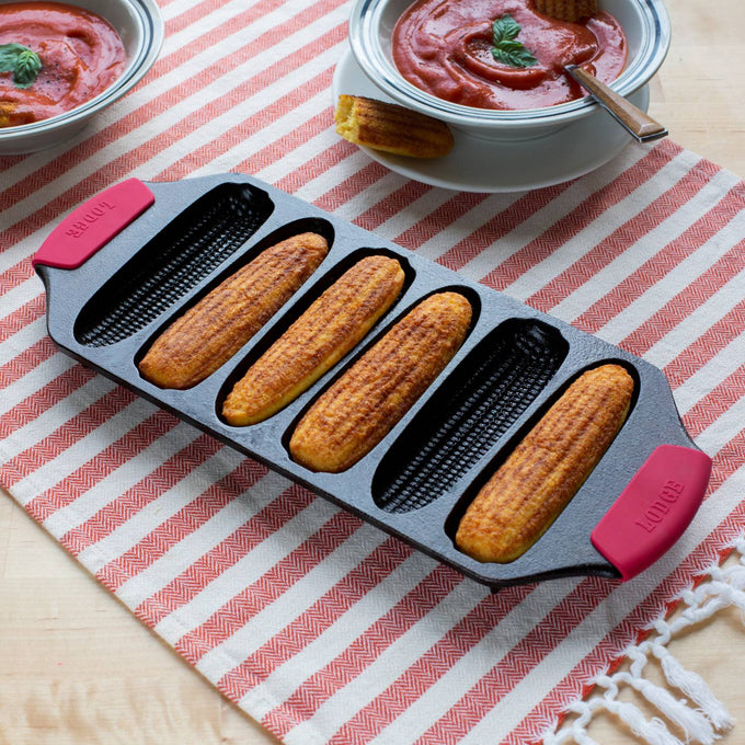 Cast iron pan with six crispy cornsticks, featuring red silicone handles, resting on a striped cloth next to bowls of tomato soup, ideal for Kitchen & Food, Cookware, Cast Iron, USA Made, and Best Sellers collections.