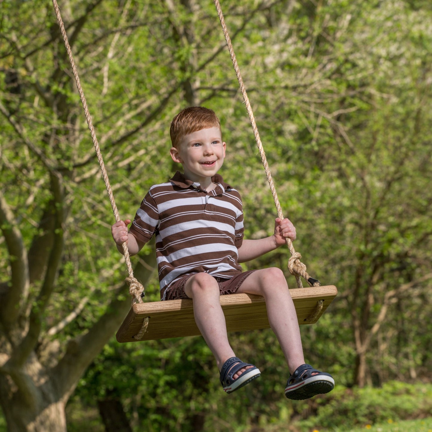 Young boy happily swinging on a handcrafted wooden rectangle-style swing seat with natural rope, outdoors among green trees, part of USA Made and Children's Toys collections.
