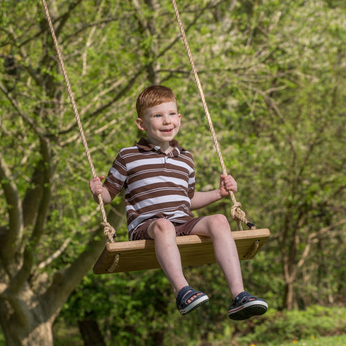 Young boy happily swinging on a handcrafted wooden rectangle-style swing seat with natural rope, outdoors among green trees, part of USA Made and Children's Toys collections.